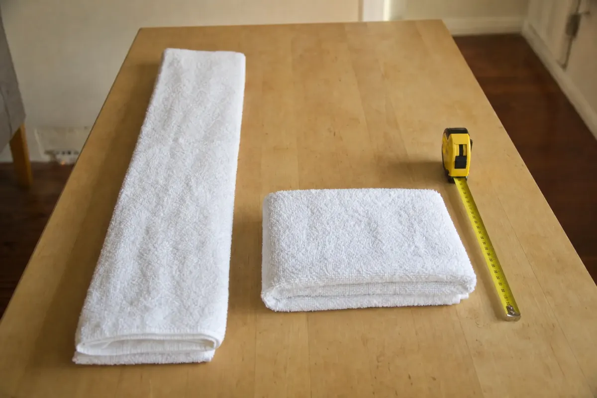 A white bath towel folded lengthwise into thirds as a long strip on a wood table, beside a second towel folded into a flat rectangle with a yellow tape measure lying along its long edge