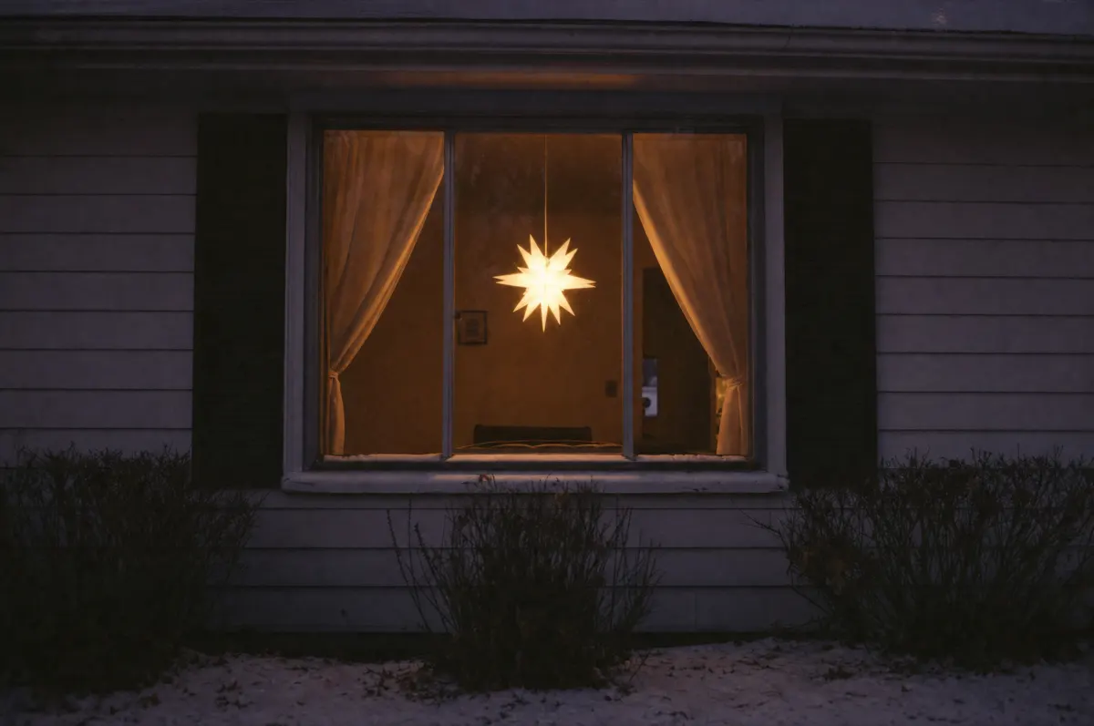 A lit white paper star centered in the front window of a small house at dusk, seen from the sidewalk, with no other decorations on the house