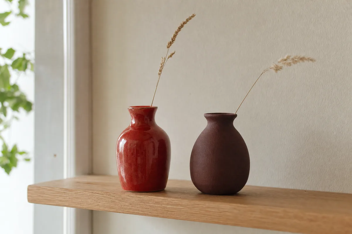 Two small vases side by side on a pale wood shelf against a cream wall, one bright cherry red and one deep brown-red, with a sprig of dried grass in each